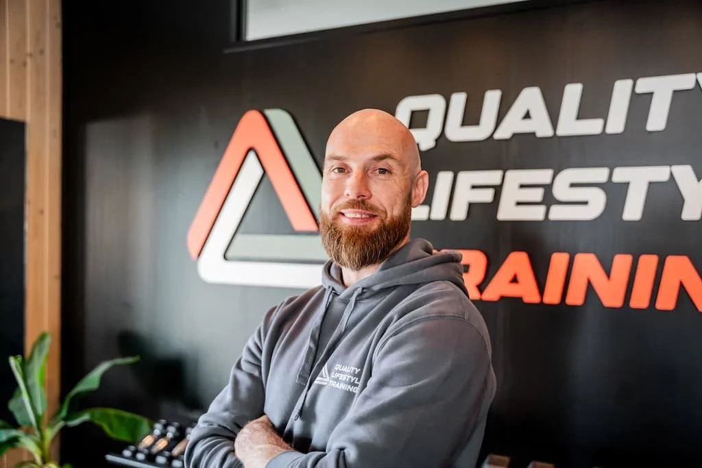 Gert-Jan, personal trainer bij Quality Lifestyle Training, poseert in de fitnessstudio voor het logo van de sportschool.